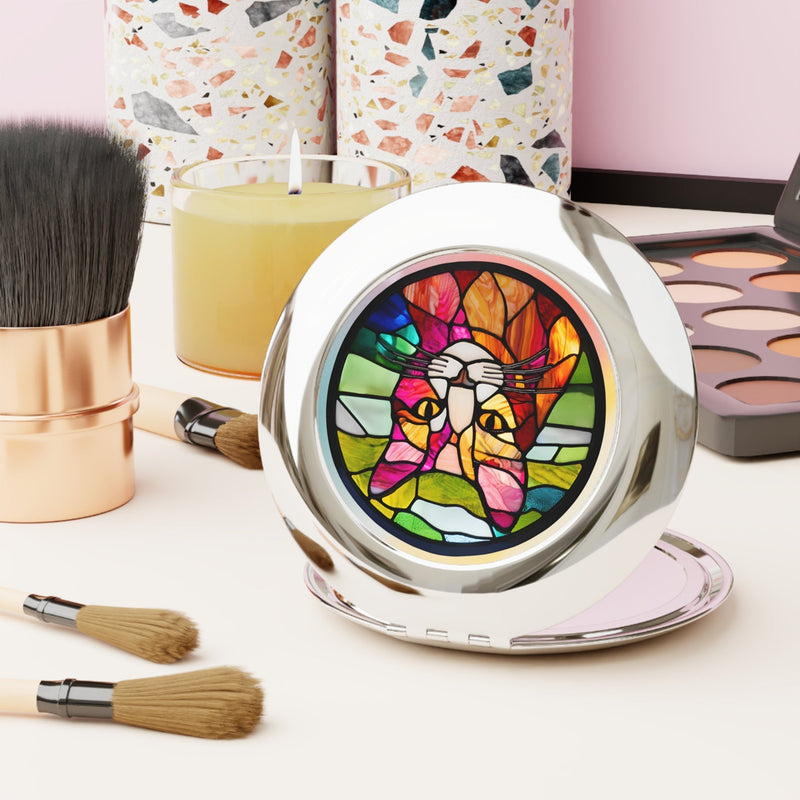 Silver women's compact mirror featuring stained-glass like image of a cat. Compact is sitting on a table with makeup brushes.